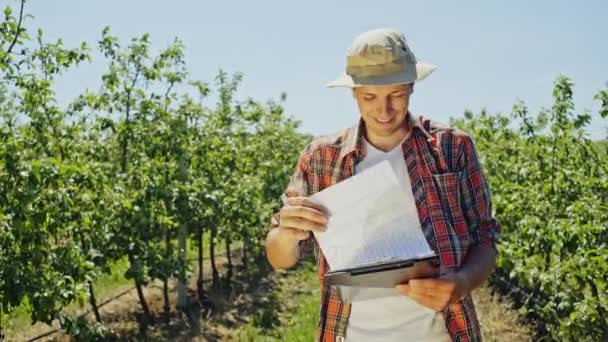 Glad agriculturist tittar igenom papper utomhus bland växter i gården plantagen på solig och blåsig dag. Ung man i hatt och röd rutig skjorta stående och tittade på från låg vinkel — Stockvideo