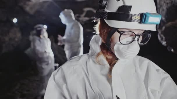 Vrouw in beschermende kleding dragen van een helm, masker en verlichte koplamp ondergronds werken met een team van collega's, het maken van notities op een clip boord in de duisternis in ondergrondse tunnel of grot. — Stockvideo