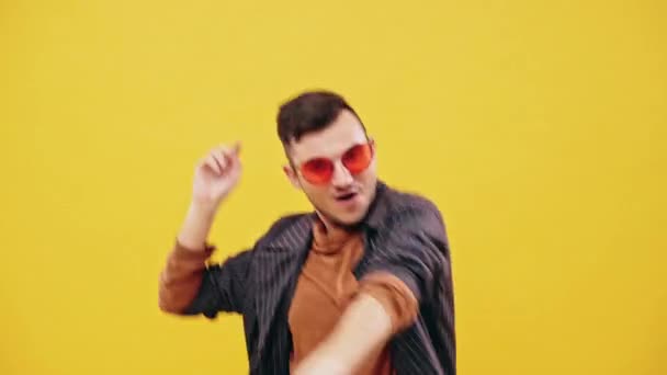 Счастливый молодой парень со стильной стрижкой, одетый в коричневую водолазку рубашку, темный пиджак и красные солнцезащитные очки танцующие в клубе или на вечеринке на обычном желтом фоне, рассматривается в половине длины . — стоковое видео