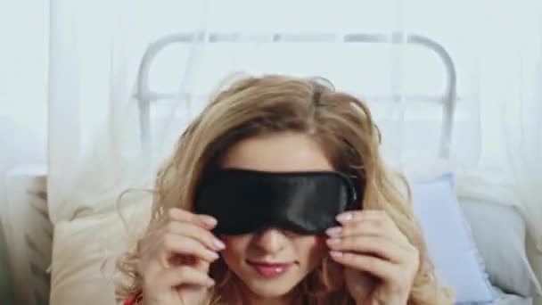 Camera following beautiful young female taking off sleep mask and blinking quickly while waking up on comfortable bed in morning — Stock Video