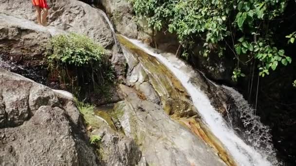 Veduta di un'alta cascata che sgorga su rocce scoscese inclinando l'altezza dell'acqua verso una giovane donna bionda in abito rosso in piedi in cima alle cascate in un paesaggio tropicale . — Video Stock