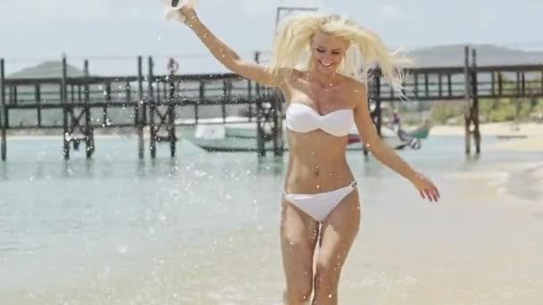 Jonge slanke blonde in witte bikini waarop tropische kust spatten in zonlicht in slow motion — Stockvideo