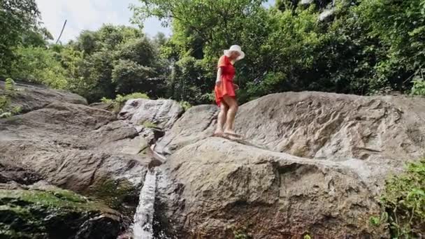 Schöne Mädchen in einem roten Kleid an einem Wasserfall in einem tropischen Wald bei sommerlichem heißem Wetter. — Stockvideo