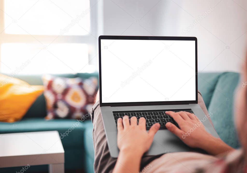 Man using, working on laptop with blank screen while lying on sofa in living room. mockup template