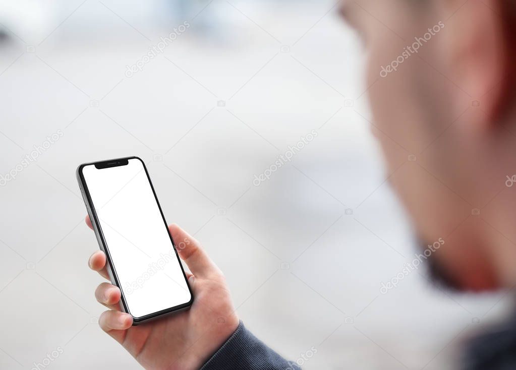 Man holding the black smartphone with big white blank screen mockup, modern frame less design, isolated on blurred background. View from above the shoulder
