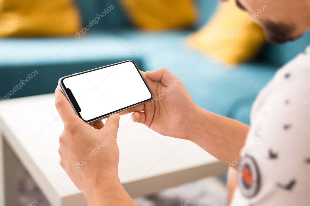 Man hand holding the black smartphone with big blank screen and modern frame less design in home interior, living room - isolated on white background horizontal position