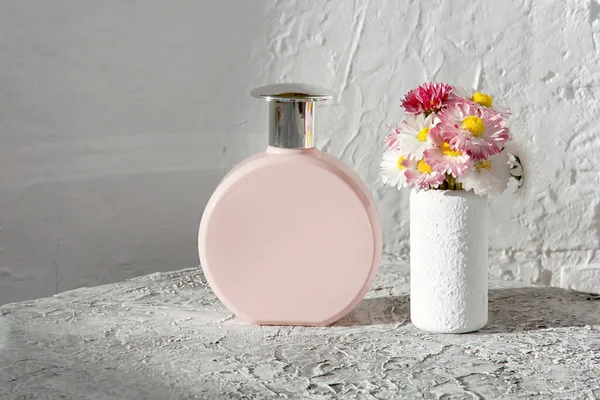 perfume in a pink powder bottle and daisies on a light texture background in defocus with blur