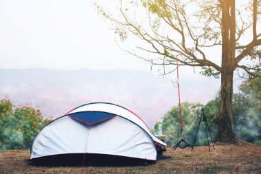 Çimlerin üzerinde ağaç ve yeşil dağlı doğa kampı çadırı. Su şişesi ve fotoğraf için tripodu olan orman ulusal parkında tatil pikniği için sis, tatil için seyahat ve sıcak güneş ışığı.