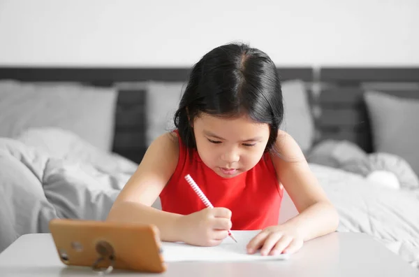 アジアの子供や子供の女の子は 思考や人々が自宅から学び 学校に戻ってテーブルの上のスマートフォンや携帯電話でオンラインで勉強し 赤いドレスを着て紙の上で読み書きや宿題を行う — ストック写真