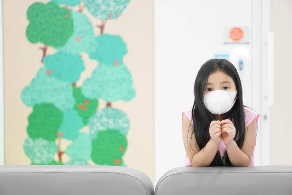 Asian child or kid girl wear N95 white face mask to close mouth nose with cold cough sick and sneeze flu to sit on sofa with social distancing for protect coronavirus or air dust at hospital or school