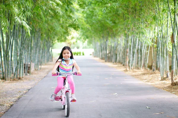 Asian child smile or kid girl wear face mask under chin and cycling fun on bike or people bicycle on public park with green garden and bamboo tree for sport exercise to healthy on summer relax holiday