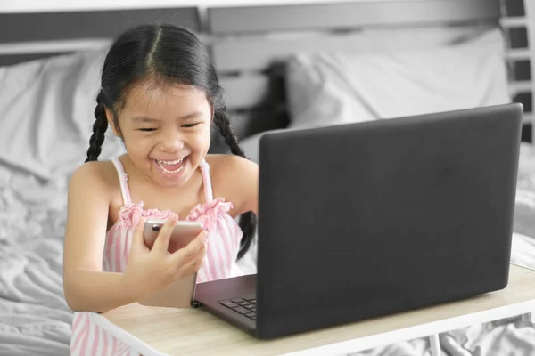Asian child student or kid girl happy smiling to shopping by smartphone or study online and work from home on computer notebook or play laptop for primary education school and stay safe in bedroom
