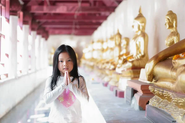 Asian child cute hand in hand or kid girl pay obeisance or respect and pray or raise with gold buddha statue and wear white shirt at temple for peace and buddhist or religious ceremony to happy smile