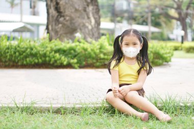 Asyalı çocuk ya da çocuk N95 kağıt maskesi takıyor ve soğuk öksürükle burun üstü hasta ve PM2.5 tozu veya grip virüsü ile bakterileri ağaç bahçesi ve şehir parkında duman havasından koruyor.