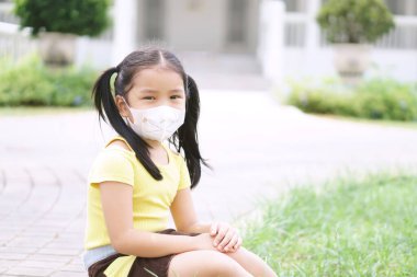 Asyalı çocuk ya da kız çocuğu N95 kağıt maskesi takıyor ve soğuk öksürükle hasta oluyor ve PM2.5 tozu veya grip virüsü ile bakterileri bahçe ve şehir parkında duman havasından koruyor.