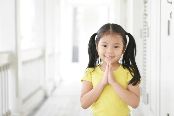 Asian child smiling hand in hand or kid girl pay obeisance and pray or hello welcome and thank you with wear yellow shirt in morning at church or temple for peace on white house receptionist at hotel