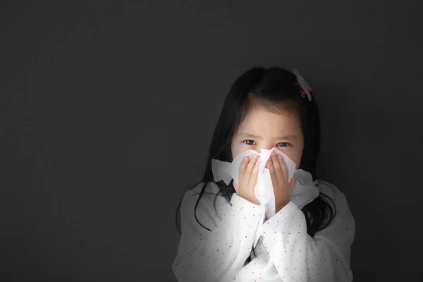 Asian child or kid girl sick and sad with sneezing on nose and cold cough on tissue paper because influenza and weak or virus bacteria from dust weather or smoke at school for medical on dark or black