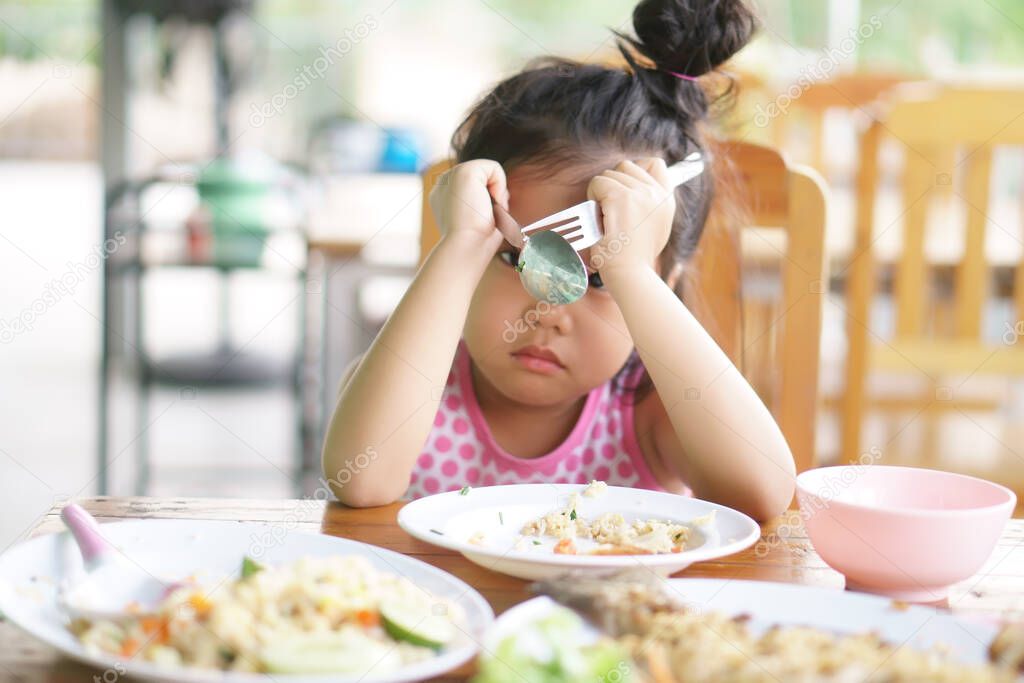 Asian child or kid girl anorexia or sad and bored food or boredom with sleepy vacant and prop up or hand to head with headache on wood table for breakfast or lunch eating at restaurant on preschool