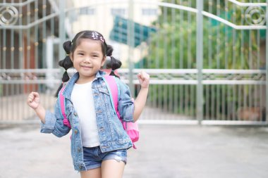 Gülümseyen Asyalı çocuk ya da kız çocuğu ve mutlu bir öğrenci omuz çantası ve güçlü sağlıklı ve kendine güvenen bir avuç dolusu insan okula ya da okula ya da yaz eğitimine ya da öğrenmeye giden anaokuluna gitsin.