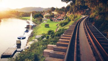 Tren ve fıskiyeler için doğal peyzaj rayları ve nehir üzerindeki yeşil çimen çayırı tatil beldesi veya turist seyahati için otel Tayland 'daki Kanchanaburi' de dinlenme tatili ve sıcak güneş ışığı vintage