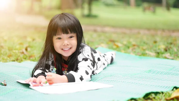Asian child cute or kid girl learning for coloring or sleep drawing paint on green mat and nature meadow at autumn winter garden with happy smile white teeth and preschool education on holiday relax