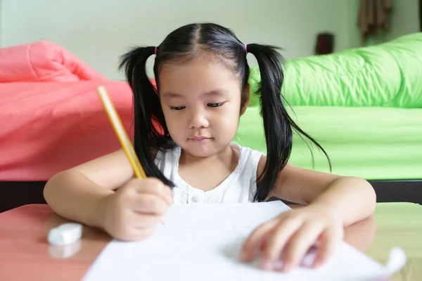 アジア人の子供はかわいいか子供の女の子は勤勉なレビューを笑って宿題や幸せな学習や訓練を行うことを楽しむ白い紙や保育園で本や家のベッドルームで木製のテーブルの上に書く — ストック写真