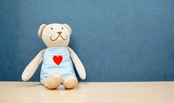 brown teddy bear doll and smile with red heart on shirt sit on table or desk and blue wall background for gift on love valentine at kindergarten or nursery and home with space