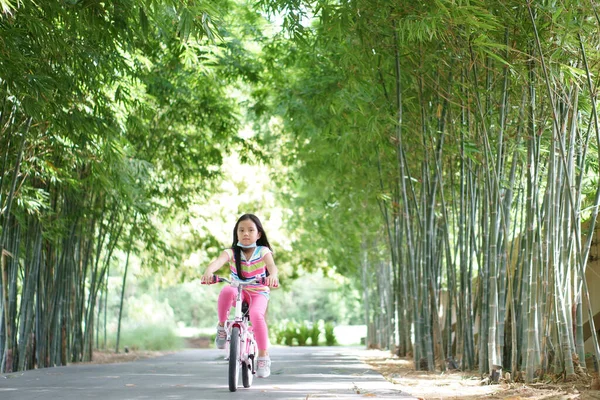 Asian child smile or kid girl wear face mask under chin and cycling fun on bike or people riding bicycle on public park and green garden or bamboo tree for sport exercise to healthy on summer holiday