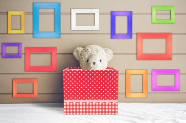 Beyaz yatağın üzerinde kırmızı hediye kutusundaki eski oyuncak ayı oyuncağı. Yatak başlığında çok renkli boş resim çerçevesi ve kahverengi duvar arkası hediye ve sürpriz için. Ya da doğum günü partisi için.