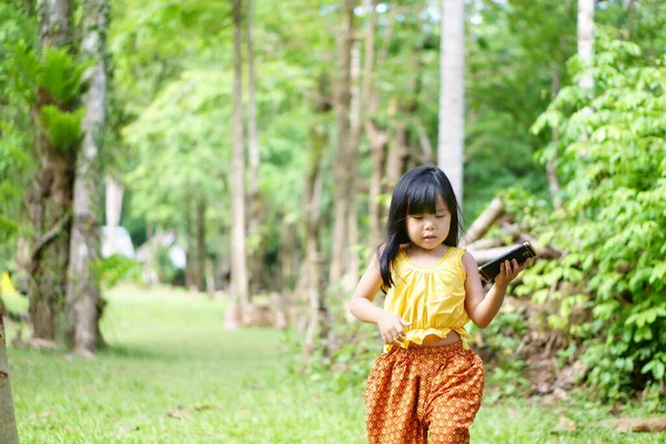 stock image Asian children cute or kid girl wear traditional top or sleeveless shirt and walk on nature jungle or tree garden with green grass for holiday relax and hold smartphone for communication or nomophobia