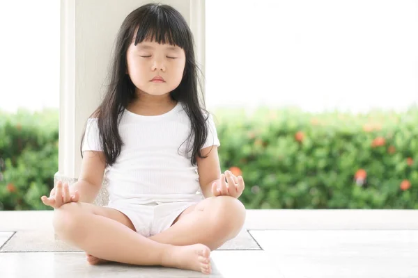 Asian children cute or kid girl sit for close eye meditation or yoga and for peace and relax or recreation in garden pavilion at temple or church and wearing white dress with nature sunlight and space