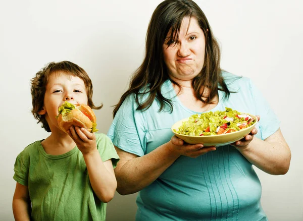 fat woman holding salad and little cute boy with hamburger teasing, real family on white