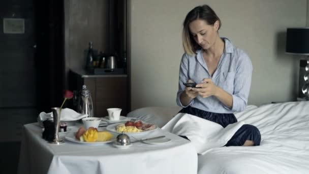 魅力的な実業家のテキスト メッセージのスマート フォンとホテルの部屋で朝食を食べる — ストック動画