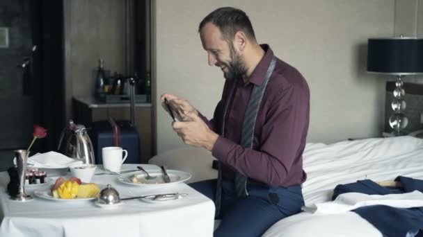 電話でゲームをホテルで朝食中に遊ぶ若い 幸せなビジネスマン — ストック動画