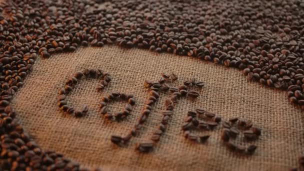Cartas con granos de café tostados. Inscripción de la palabra "café" hecha de frijoles en la textura de la bolsa de yute. Movimiento de la cámara con rotación  . — Vídeo de stock