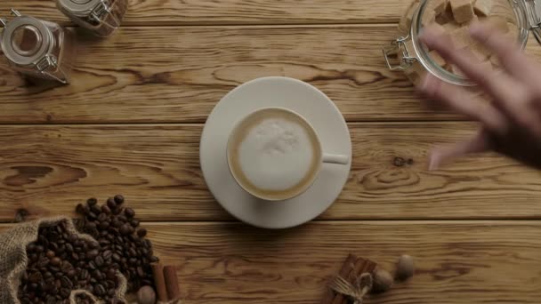 Mit der Hand braune Zuckerwürfel hinzufügen und mit einem Löffel in die Cappuccino-Kaffeetasse rühren. Holztisch — Stockvideo