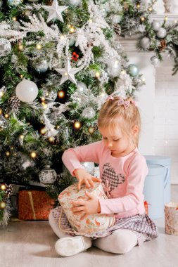 Küçük bir kız Noel ağacının altında içinde Noel hediyeleri olan bir hediye kutusu açıyor.