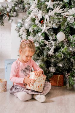 Küçük bir kız Noel ağacının altında içinde Noel hediyeleri olan bir hediye kutusu açıyor.
