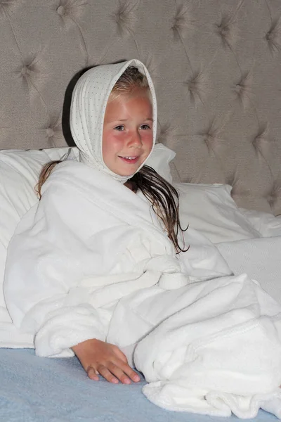 a girl in a white robe and a white scarf on her head with happy face is sitting on the bed