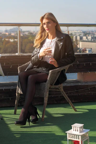 a beautiful middle aged Russian woman is sitting in a wicker chair on the roof with a glass of champagne in her hands and a view of the autumn city in the bright light of the warm sunset