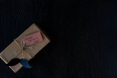 Koyu ahşap bir arka planda el işi kağıttan yapılmış hediye paketi ve kağıttan yapılmış bir bıyık. Babalar Günü kavramı, Anavatan Günü 'nün koruyucusu.