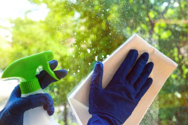 Pencereleri beyaz bir bezle yıkamak mavi eldivenli bir el ve temizlik spreyi ile. Pencerenin dışında yemyeşil ve parlak güneş ışığı var.