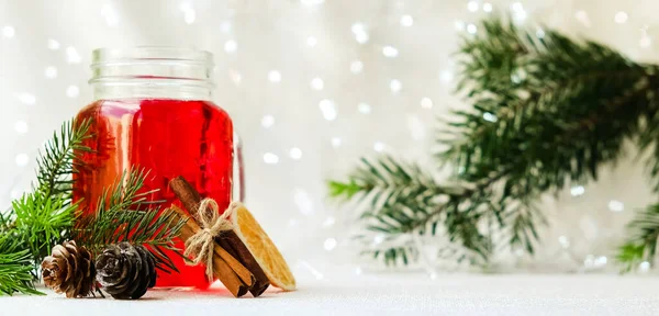 a jar with a handle with red mulled wine on a white background with a garland. Next to a branch of spruce and cinnamon and orange. The concept of Christmas