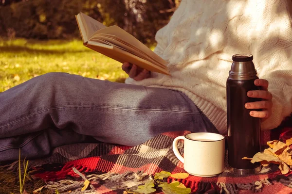 在秋天的森林里 一个美丽的女孩正在看书 上面涂满了温暖的白 一个女人坐在秋天森林里的一棵树旁 手里拿着一本书和一杯酒 手里拿着一杯热饮 正在看书的女孩 免版税图库照片
