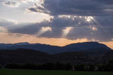 Gün batımında kara bulutların arasından yükselen güneş ışınları. Önünde bir tarla, arkasında dağlar var. Görüntü yatay. Peyzaj kavramı.
