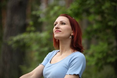 Dışarıdaki doğal ortamda duran kızıl saçlı kadının portresi.