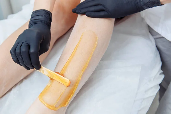Siyah lateks eldivenli bir el tıraş edilmemiş bir bacağa sarı balmumu sürer..