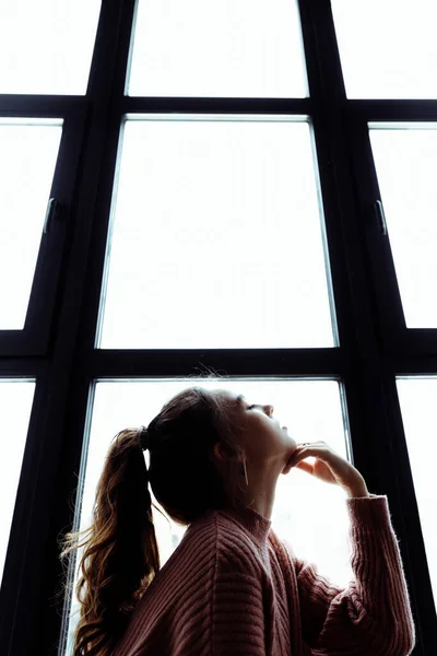 La chica está sentada frente a la ventana. El pelo de las niñas se tira hacia atrás en una cola de caballo — Foto de Stock