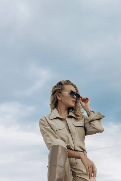 Mavi bulutlu gökyüzüne karşı şık bir tulum giymiş sarışın bir kadın başka tarafa bakıyor..