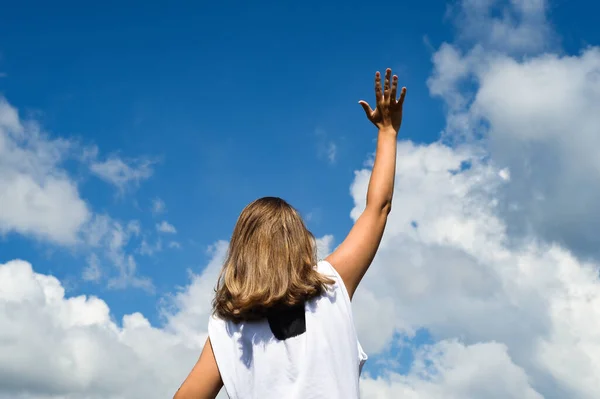 A woman stands with her back to the photographer in a white T-shirt and against a blue sky with clouds. Reaches her hand to the sky.